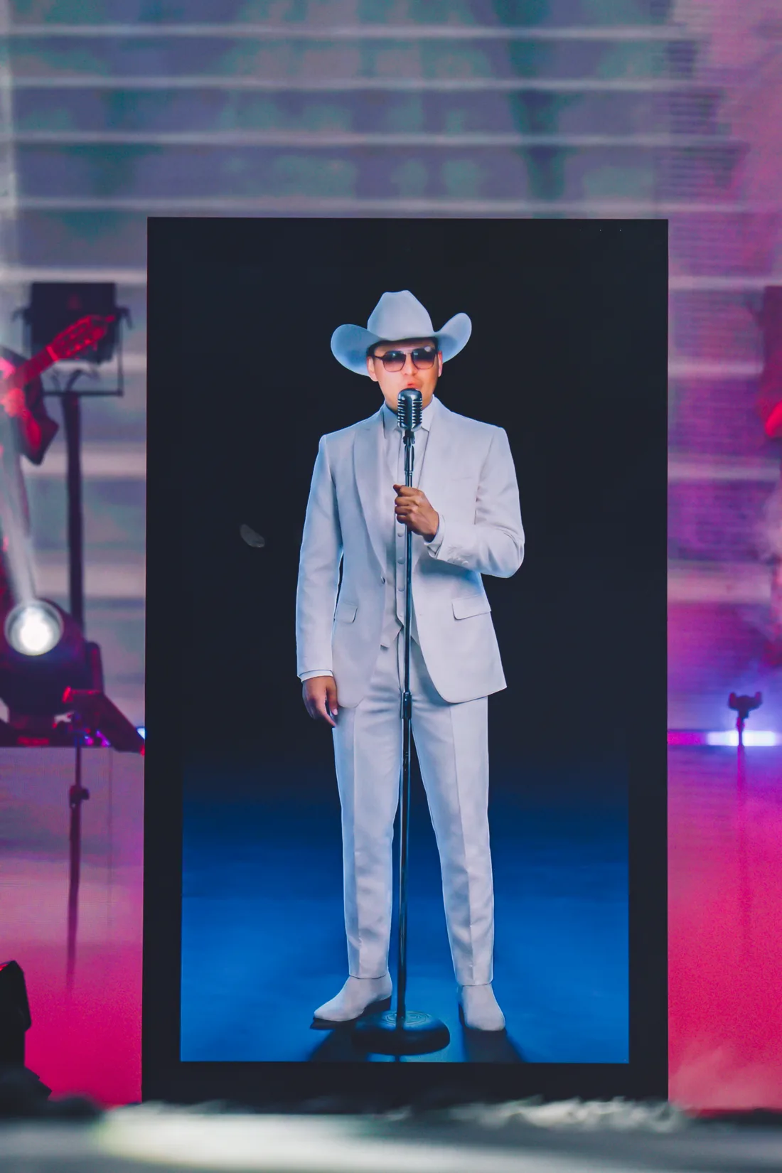 Plano cercano del avatar hiperrealista de Yeison Jiménez con traje blanco, sombrero y micrófono, generado con inteligencia artificial, dentro del marco vertical del escenario